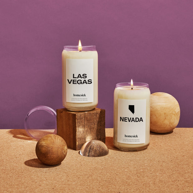 Two Homesick Candles shot on a corkboard surface with a deep purple background. The two shown homesick candles are the Las Vegas and Nevada candles.