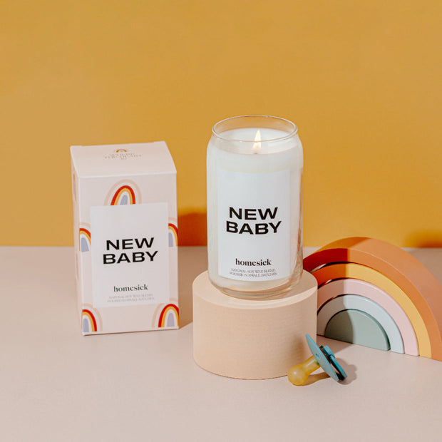A stylized studio shot of the New Baby candle displayed on a cream rounded pedestal. To the left is the box packaging for the candle with a wooden rainbow toy on the right. 