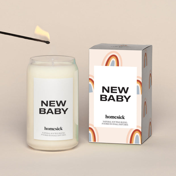 A lit New Baby Homesick candle displayed next to its boxed packaging on a dark cream background.