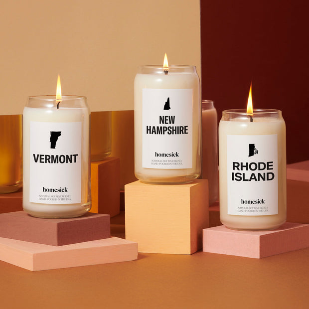 A group shot of the North East inspired candles available at Homesick. They each are displayed on top of square and rectangular pedestals in a warm orange and brown studio environment,