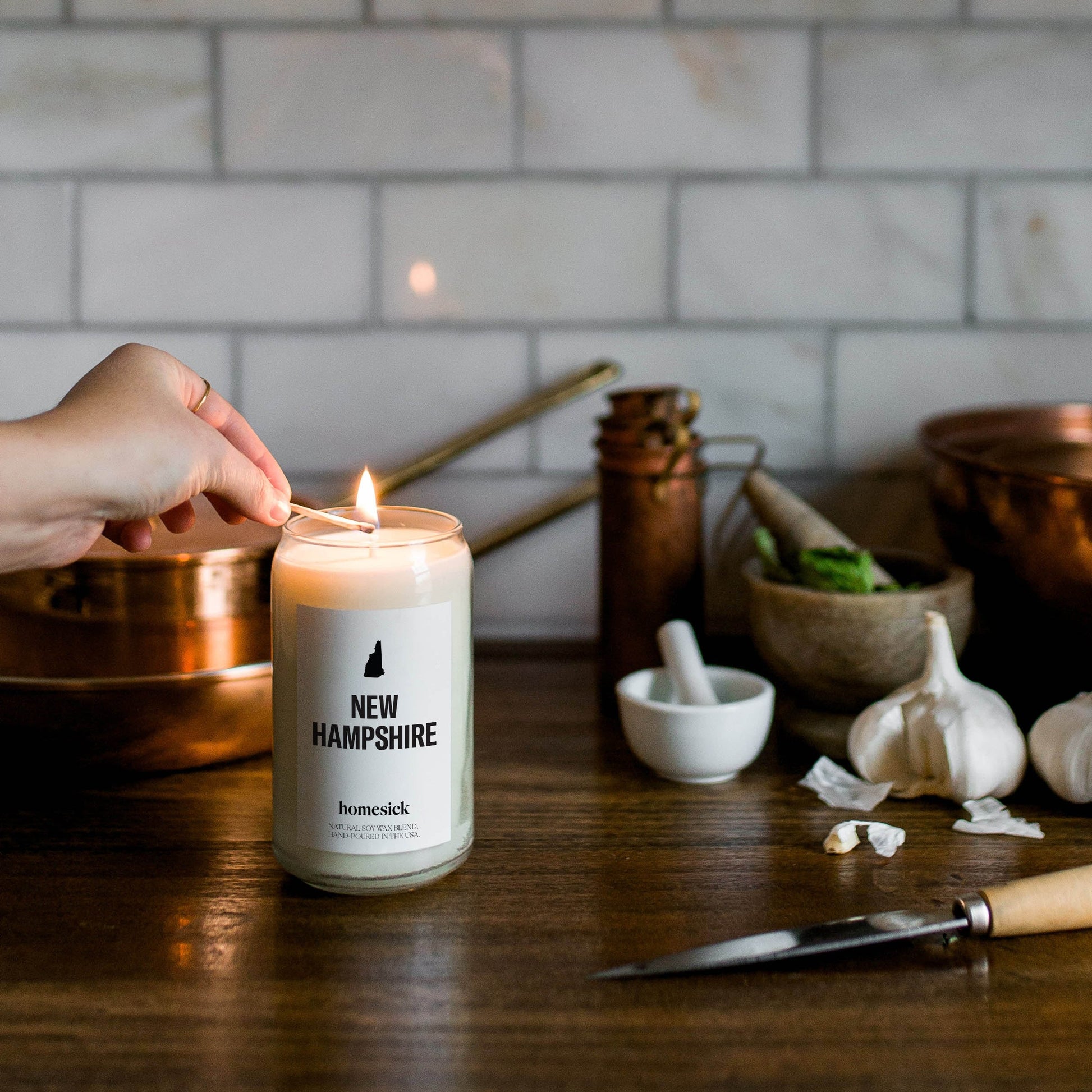 A hand going to light the New Hampshire candle with a match inside a kitchen that has various prep items.