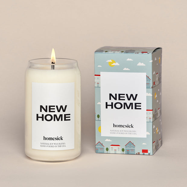 A lit New Home Homesick candle displayed next to its boxed packaging on a dark cream background.