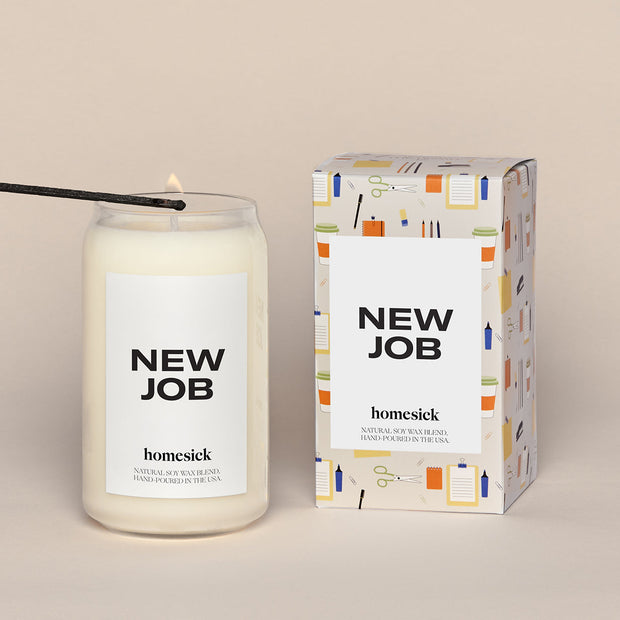 A lit New Job Homesick candle displayed next to its boxed packaging on a dark cream background.