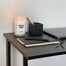 A shot of the corner of a black desk situated next to a white wall. On the desk is a lit New Job candle, a black notebook, laptop and black pen holder.