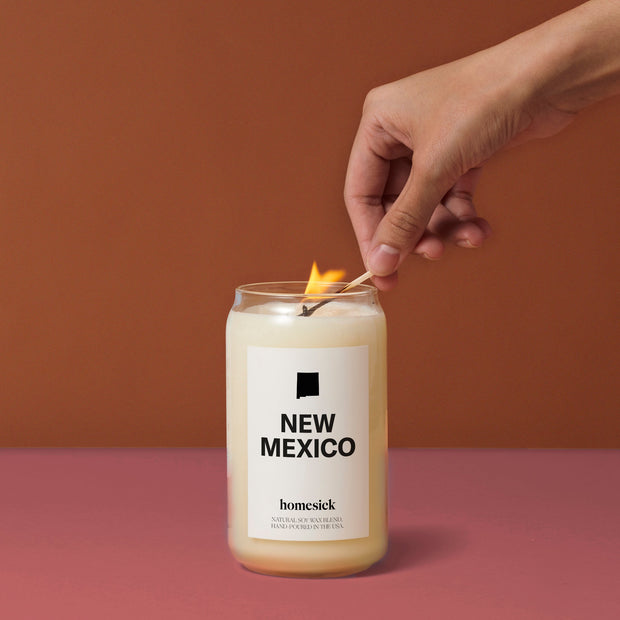 A New Mexico candle on a dark amber surface with a darker background. A hand is coming in from the top right corner with a match to light the candle.