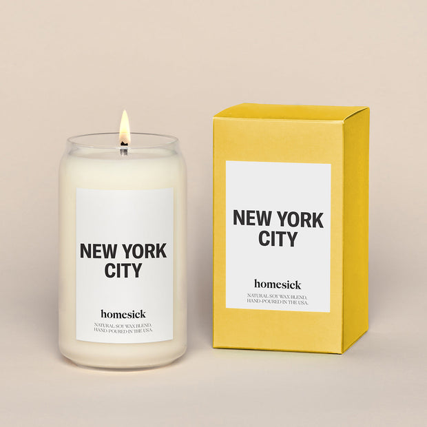 A lit New York City Homesick candle displayed next to its boxed packaging on a dark cream background.