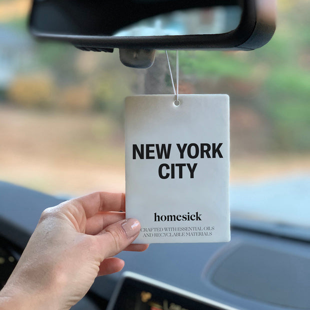 A hand touching the New York City Car Freshener that is hanging from their rearview mirror. The background is blurred.