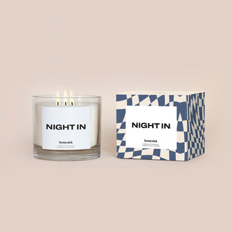 A lit 3-Wick Night In Homesick candle displayed next to its boxed packaging on a dark cream background.