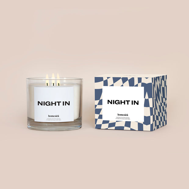 A lit 3-Wick Night In Homesick candle displayed next to its boxed packaging on a dark cream background.