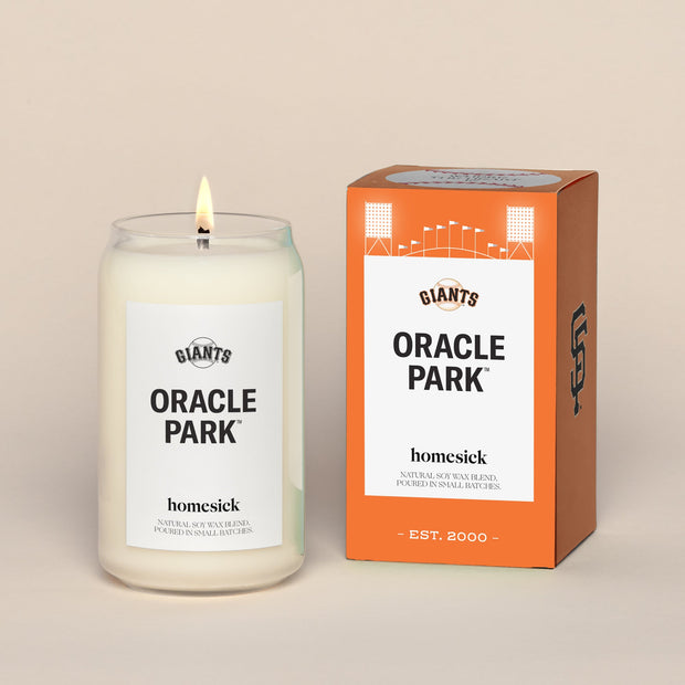 A lit Oracle Park Homesick candle displayed next to its boxed packaging on a dark cream background.