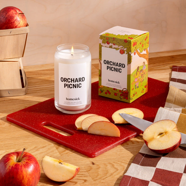 A stylized shot of the Orchard Picnic candle and its boxed packaging on a red cutting board that sits on top of a wooden surface. There are apple slices around the candle.