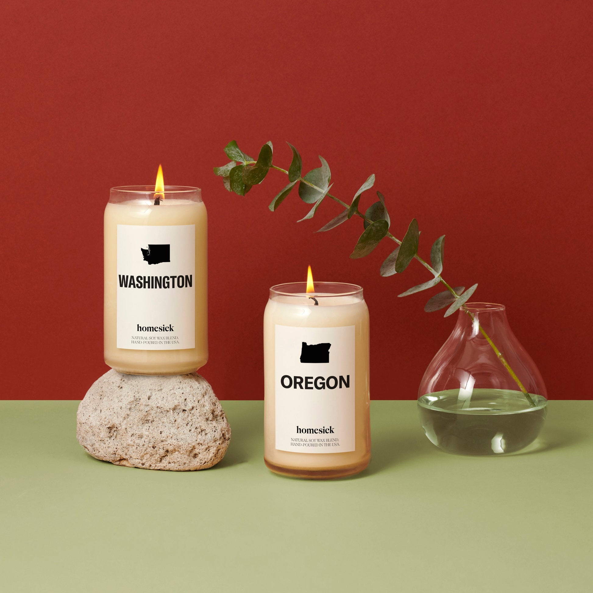 Two Northwest inspired Homesick candles on a mint green surface with a red background. The Washington and Oregon Candles are side by side with a glass vase to the right and eucalyptus coming out the vase.
