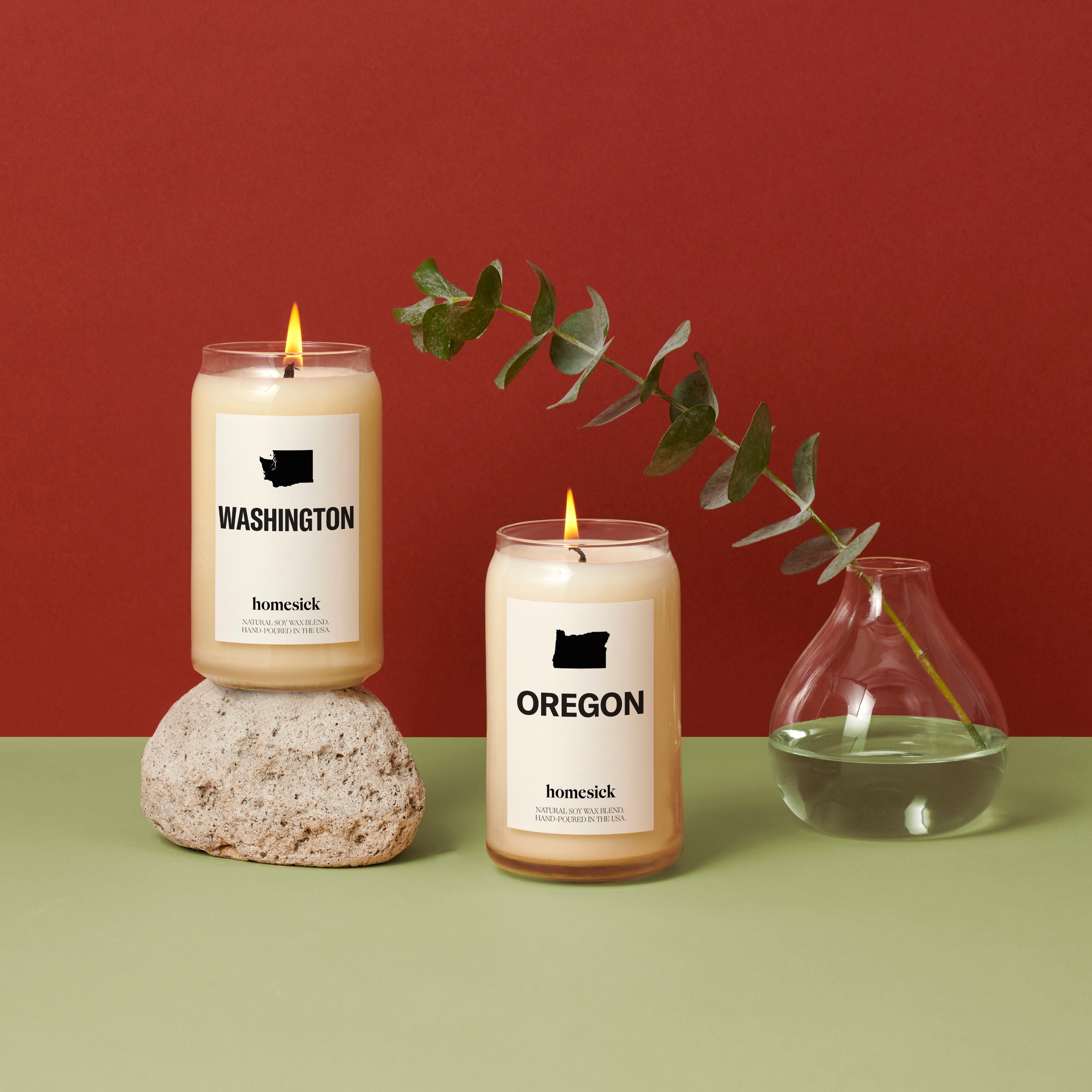 Two Northwest inspired Homesick candles on a mint green surface with a red background. The Washington and Oregon Candles are side by side with a glass vase to the right and eucalyptus coming out the vase.
