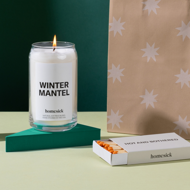 On the left is a lit Winter Mantel candle that is displayed on a dark green triangle pedestal. To the right there is a box of hot and bothered matches. Both are on a light green surface with dark background and a star gift bag poking in the side of the right side.