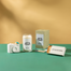 A group shot of various Homesick products on a green surface with  pale mustard yellow background. Left to right is a camera match holder, let's toast candle, lets toast packaging, and hot & bothered matches.