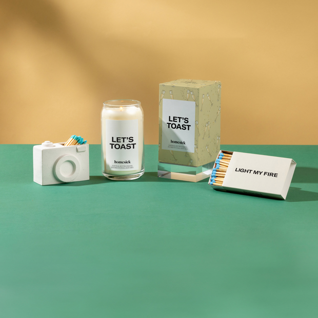 A group product shot of various Homesick products on a green surface and yellow background. Left to right are the camera match holder,let's toast candle, let's toast packaging and the light my fire matches.