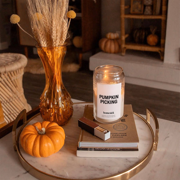 A lifestyle image of the Pumpkin Picking candle shot in a cozy fall apartment. The candle is displayed on a couple of books that also have a box of matches next to them.