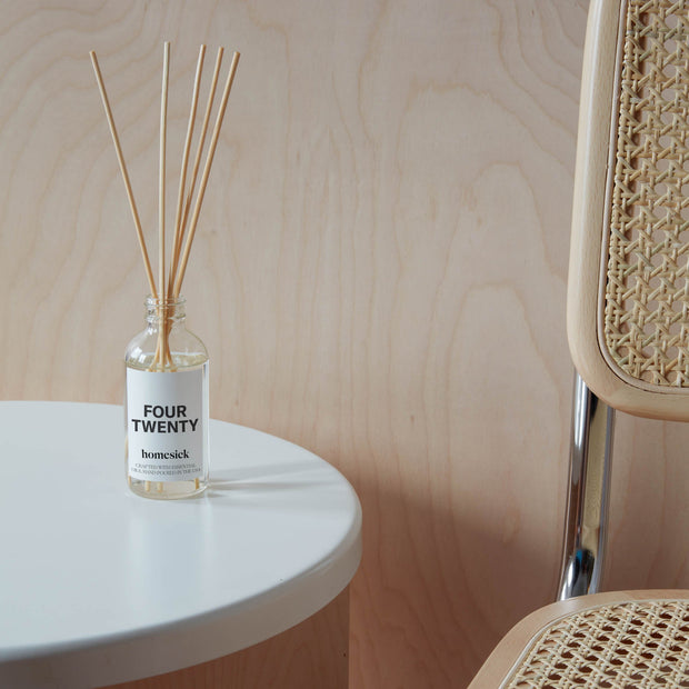 A lifestyle shot of the 420 Reed Diffuser on a white side table. One can see the wooden background a wick chair next to it.