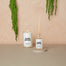 A simple styled shot of the Home Office Reed Diffuser and the Home Office Candle to its left. They are shot on a dark sand color surface and a light wood background. There is hanging moss coming down the background.
