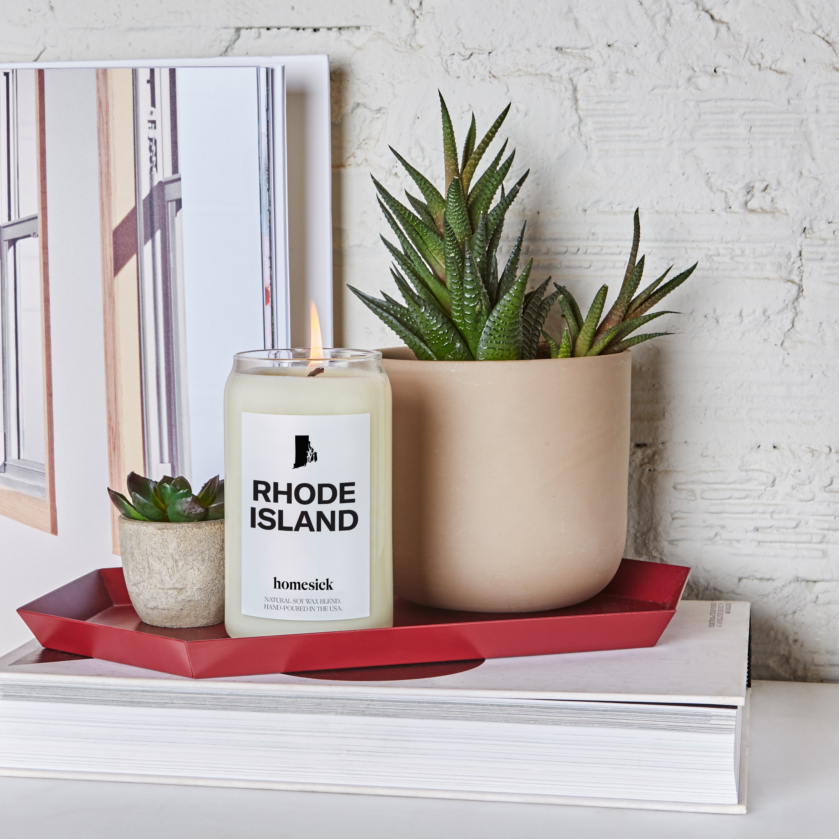 A lifestyle image of the Rhode Island candle inside a red decor try with two succulent plants surrounding it. The tray is displayed on a big coffee table book with a white brick backgroud.