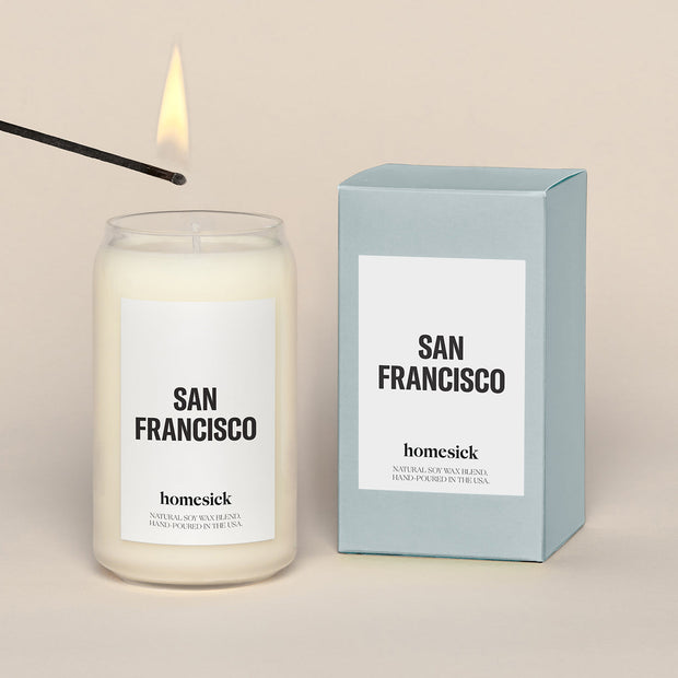 A lit San Francisco Homesick candle displayed next to its boxed packaging on a dark cream background.