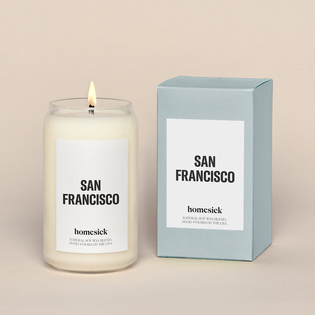 A lit San Francisco Homesick candle displayed next to its boxed packaging on a dark cream background.