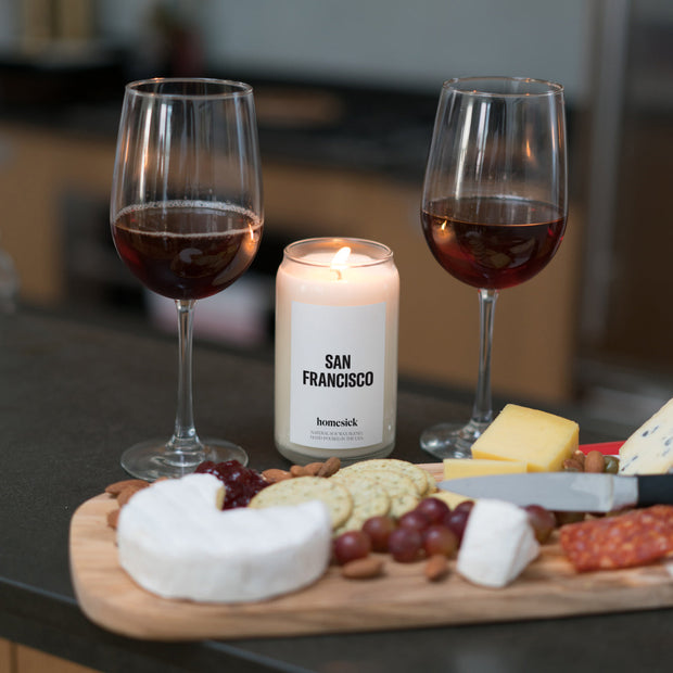 A San Francisco candle displayed in between two glasses of red wine behind a charcuterie board.