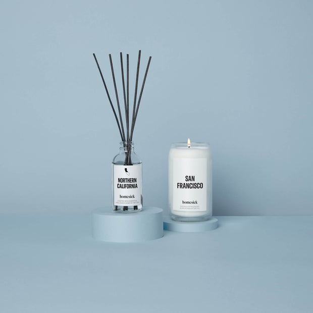 The Northern California reed diffuser next to the San Francisco candle that are in a light blue studio environment.