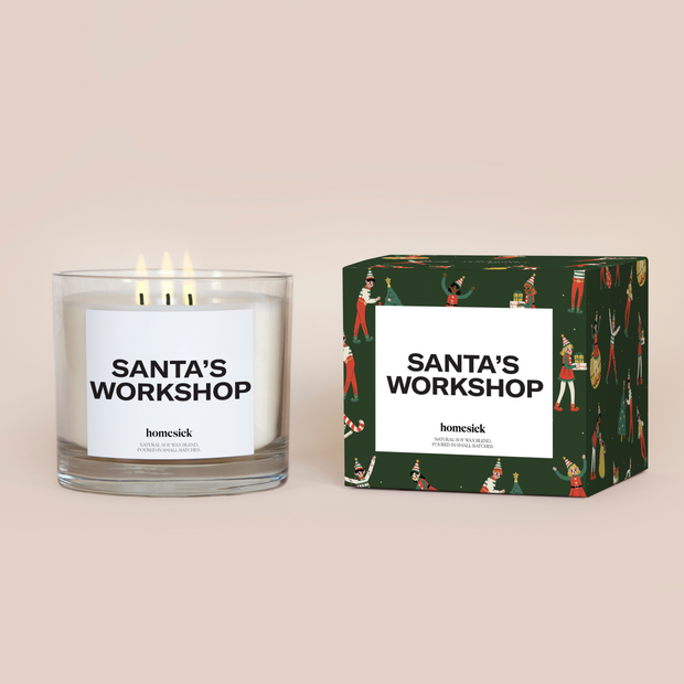 A lit 3-Wick Santa's Workshop Homesick candle displayed next to its boxed packaging on a dark cream background.