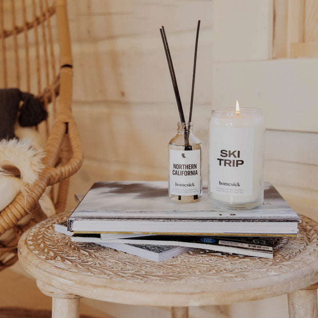 The Ski Trip Candle next to the Northern California reed diffuser. Both are displayed on a stack of magazines. 