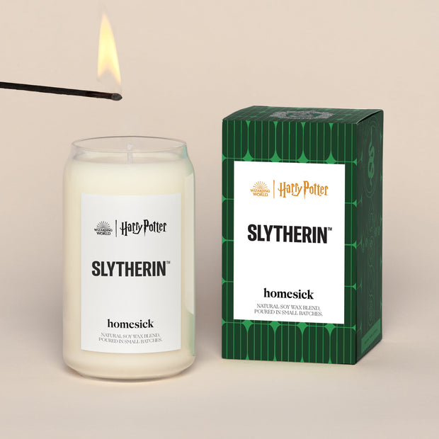 A lit SlytherinHomesick candle displayed next to its boxed packaging on a dark cream background.