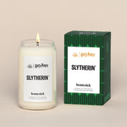 A lit SlytherinHomesick candle displayed next to its boxed packaging on a dark cream background.