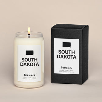 A lit South Dakota Homesick candle displayed next to its boxed packaging on a dark cream background.