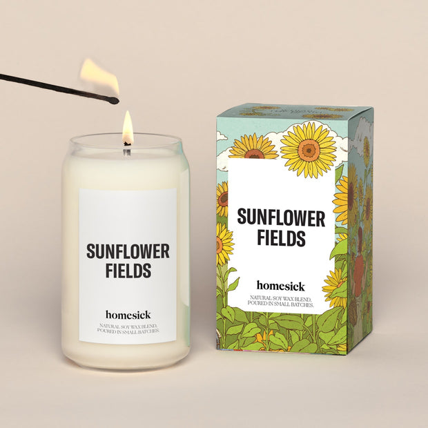 A lit Sunflower Fields Homesick candle displayed next to its boxed packaging on a dark cream background.