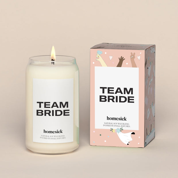 A lit Team Bride Homesick candle displayed next to its boxed packaging on a dark cream background.