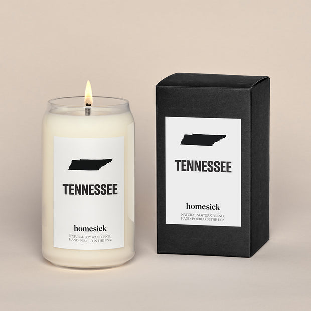 A lit Tennessee Homesick candle displayed next to its boxed packaging on a dark cream background.