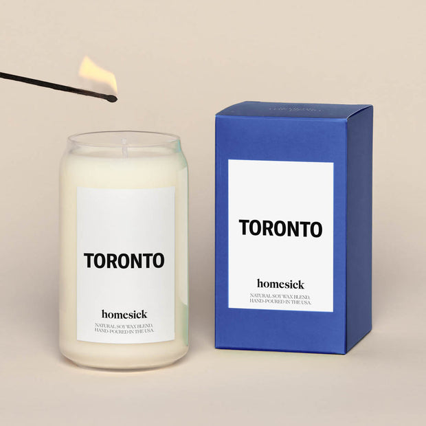 A lit Toronto Homesick candle displayed next to its boxed packaging on a dark cream background.