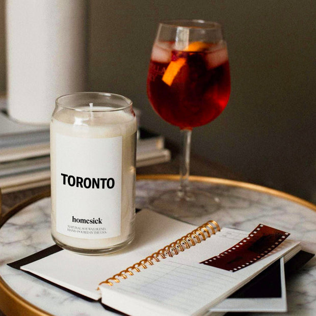 A close up of the Toronto candle on a marble side table that has a gold rim. There is an open notebook and glass of sangria also on the table.