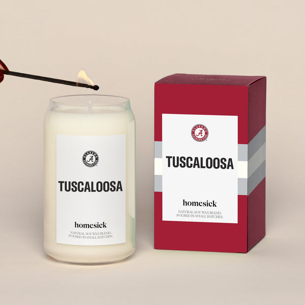 A lit Tuscaloosa Homesick candle displayed next to its boxed packaging on a dark cream background.