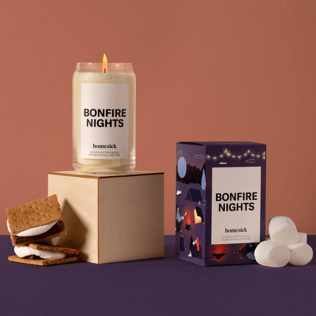 On the left, there is a lit Bonfire Nights Candle on top of a cream box. Next to it, is the packaging for this candle. Both are on top of a purple surface with an orange background. There are s'mores props around the candles.