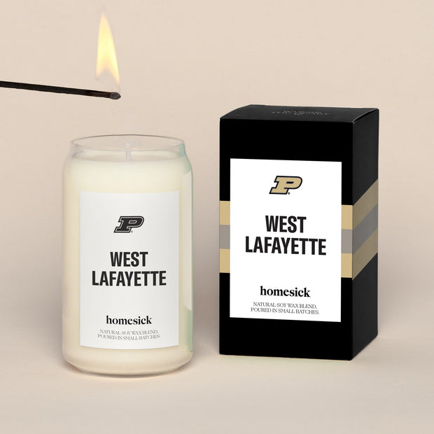 A lit West Lafayette Homesick candle displayed next to its boxed packaging on a dark cream background.