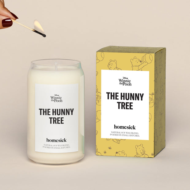 A lit The Hunny Tree Homesick candle displayed next to its boxed packaging on a dark cream background.