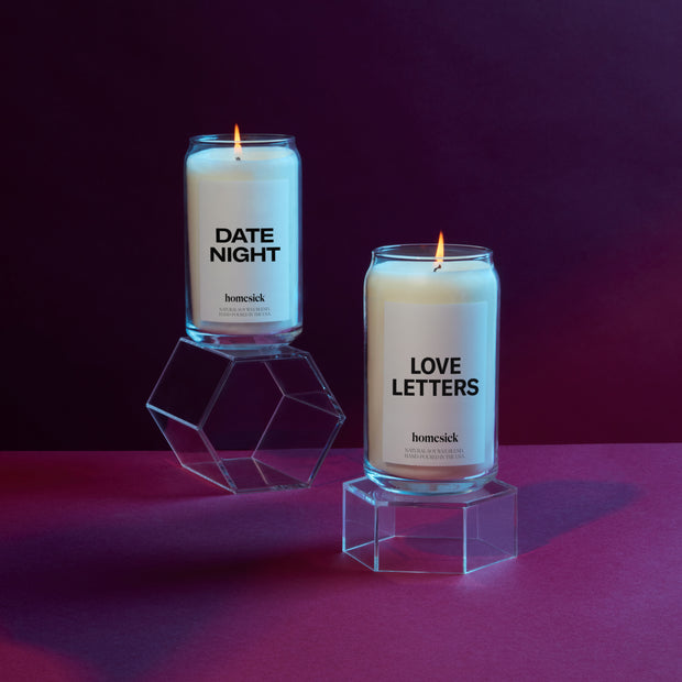 Two Homesick candles on acrylic pedestals that are on a deep purple surface with a deeper purple background. The candles on the pedestals are Date Night and Love Letters.