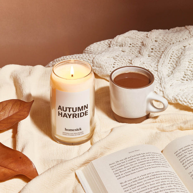 A cozy shot a lit Autumn Hayride candle, cup of coffee and a open book. They are on top of a cream blanket and can see a light brown background.. 